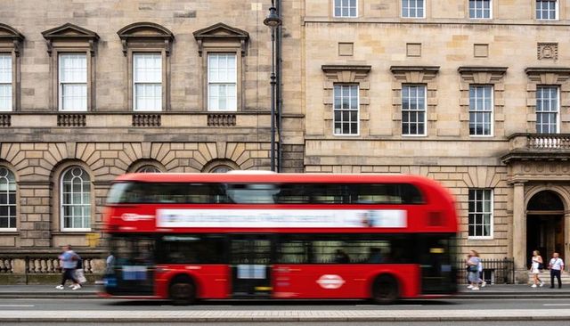 Red double-decker bus passing georgian stone building with motion blur and pedestrians