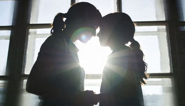 Silhouetted girls touching foreheads holding hands by sunlit window with lens flare