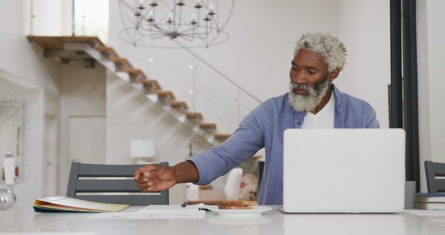 Senior African American man working from home, using laptop and reviewing documents in modern kitchen. Ideal for articles on remote work, senior professionals, or home office setups.