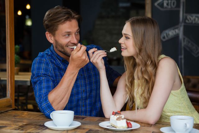 Young couple sharing a dessert while sitting at a wooden table in a cozy cafe. Both are smiling and enjoying each other's company, creating a warm and romantic atmosphere. Ideal for use in advertisements for cafes, dating apps, lifestyle blogs, or social media posts promoting relationships and leisure activities.