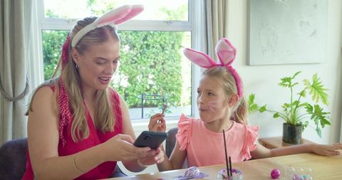 Mother and daughter painting easter eggs and crafting with bunny ears by sunlit window
