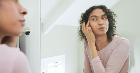 Non-binary person adjusting curls at vanity mirror in pastel minimalist bathroom