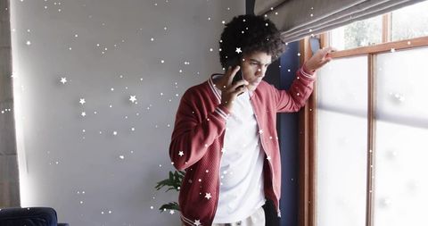 Young man talking on phone while adjusting blinds by sunlit window in modern living room