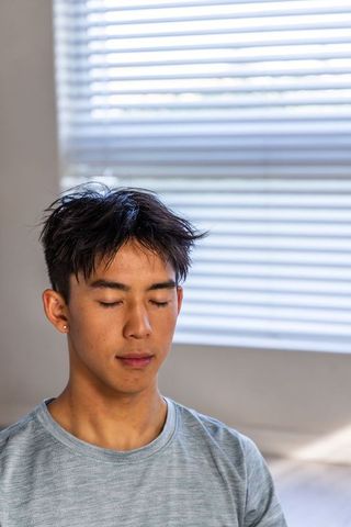 Young asian man resting with eyes closed by sunlit window blinds minimal calm portrait