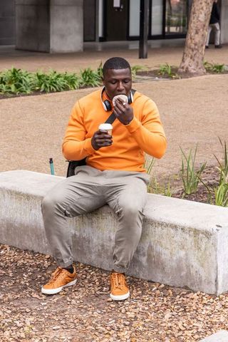 African american student enjoying coffee and donut on campus bench with headphones