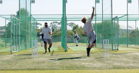 African american bowler delivering pace ball in cricket nets with teammates practicing