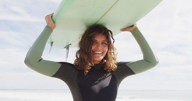 Young woman with curly hair holding surfboard over head while standing on sandy beach by ocean, wearing a wetsuit. Ideal for promoting beach vacations, summer activities, surfing gear, and healthy lifestyle campaigns.