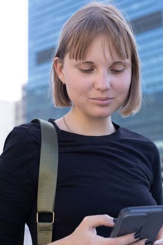 Young woman using smartphone on urban sidewalk wearing black top and olive bag strap