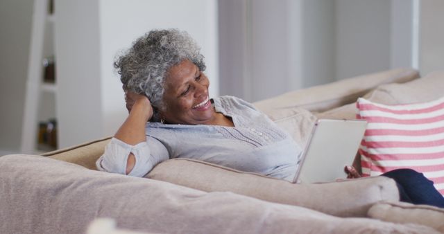 Elderly woman with gray curly hair sitting comfortably on a beige couch, using a tablet and smiling. Ideal for ads about senior living, digital literacy among the elderly, or technology-related products for seniors.