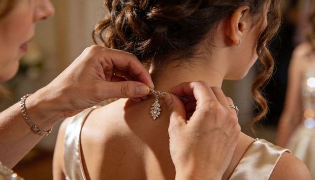 Mother fastening silver leaf necklace on bride in satin dress with elegant updo close-up