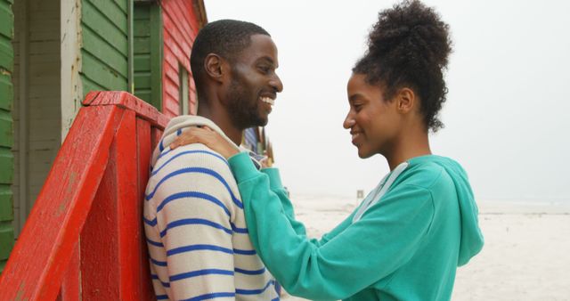 A young African American couple shares a joyful moment together by a colorful beach hut, with copy space. Their smiles and affectionate gaze suggest a romantic connection and happiness in each other's company.