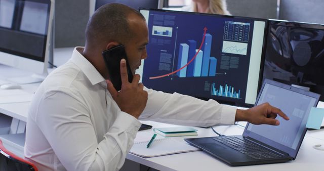 Businessman is multitasking by discussing on phone while pointing at data on laptop. Financial graphs and charts shown on computer screens. Ideal for illustrating concepts of business analysis, financial strategies, multitasking in corporate settings, and technology use in modern offices.