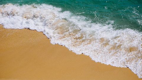 Turquoise waves crashing onto golden sand beach from above with white foam and texture