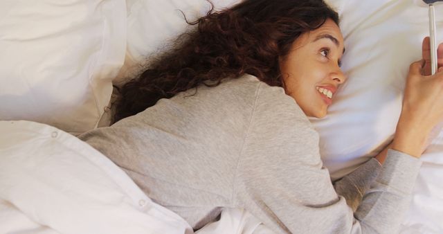 A young woman of diverse ethnicity is lying in bed, smiling as she looks at her smartphone, with copy space. Her relaxed demeanor suggests she might be enjoying a casual conversation or browsing through social media.