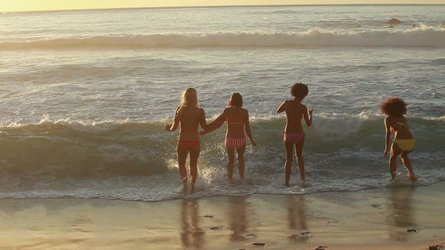 Four young women run towards the ocean, enjoying a fun and carefree moment at sunset. Ideal for themes around friendship, summer vacations, beach activities, youth, and relaxation. This video can be used in travel brochures, summer event promotions, lifestyle blogs, and advertisements depicting leisure and enjoyment.