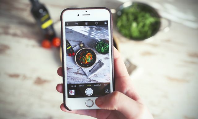 Person capturing food photo using smartphone, focusing on hand holding smartphone. Ideal for topics involving food blogging, social media content creation, smartphone photography tips, or culinary arts.