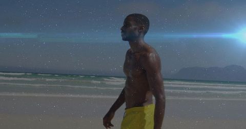 Athletic man walking sunlit beach in yellow swim trunks with cinematic lens flare