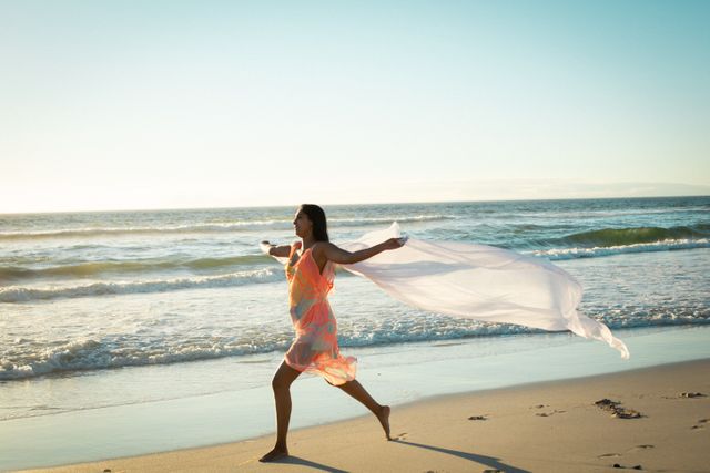 This image captures a joyful woman running along the beach at sunset, holding a flowing scarf. Perfect for use in travel brochures, lifestyle blogs, wellness websites, and advertisements promoting vacation destinations, freedom, and leisure activities.