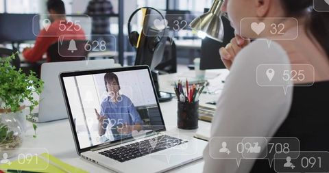 Korean presenter on laptop with social media overlays while colleague watching in open-plan office