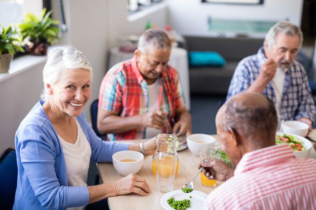 Group of elderly friends having breakfast at table in nursing home. Senior woman smiling at camera while enjoying meal with friends. Ideal for illustrating senior living, retirement homes, elderly care services, and social activities for seniors.