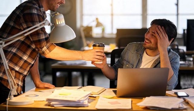 Smiling coworker handing takeaway coffee to colleague at busy open-plan office desk