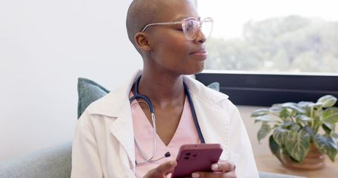 African female clinician checking smartphone while sitting in modern clinic waiting area