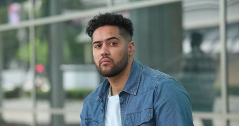 Contemplative man sitting on bench leaning by glass building wearing denim jacket