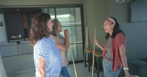 Three women laughing and chatting in modern living room holding wooden sticks casual gathering