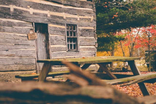 A rustic log cabin sits nestled among colorful autumn foliage. A weathered wooden picnic table is in the foreground, adding to the cozy, countryside atmosphere. Suitable for promoting rural travel destinations, outdoor adventures, nature retreats, or seasonal greeting cards.