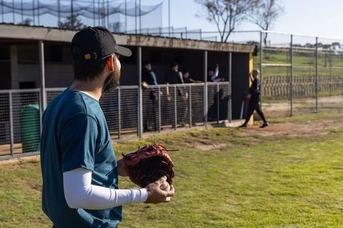 Bearded player holding glove and ball watching team in dugout at community baseball field