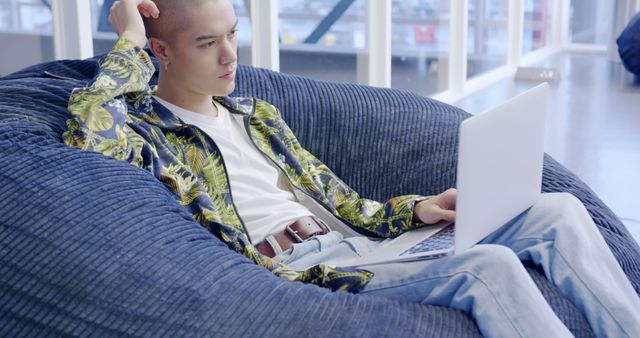 A young man is relaxing on a large blue bean bag chair while using a laptop. He appears calm and focused, dressed casually in a white t-shirt, bright patterned shirt, and jeans. The setting is contemporary, likely a coworking space or a home office. Ideal for depicting modern leisure, focusing on comfort while using technology, or illustrating relaxed work environments.