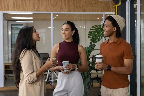 Multicultural coworkers chatting, holding coffee in modern glass-walled office break area