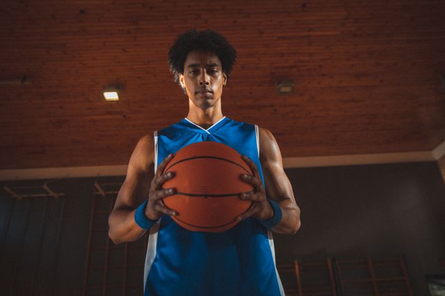 Portrait of biracial male basketball player holding basketball in two hands. basketball, team sports training at an indoor court.