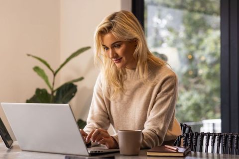 Young woman working from home on laptop with coffee in cozy sunlit modern office by window