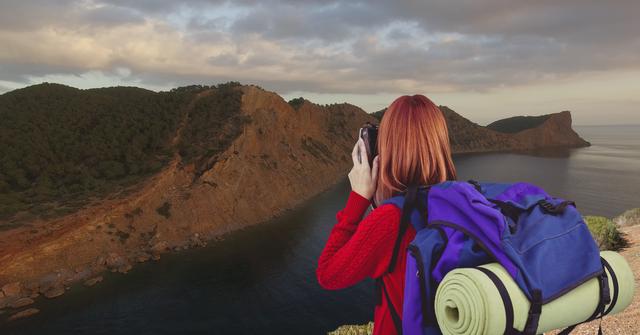 A red-haired woman with a large backpack is photographing a mountain landscape. This visual can be used to represent themes such as adventure, exploration, traveling, hiking, and scenic photography. Ideal for tourism promotions, travel blogs, and outdoor activity advertisements.