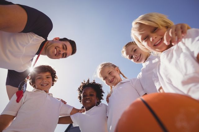 Group of diverse schoolchildren with their coach in a schoolyard, smiling and bonding over a basketball activity. Ideal for use in educational materials, sports programs, youth development campaigns, and advertisements promoting teamwork and physical education.