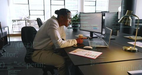 Mature professional typing on laptop in open-plan office with monitors, charts and coffee