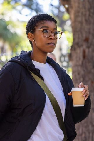 African american woman holding coffee cup in puffer jacket and glasses walking in sunlit park