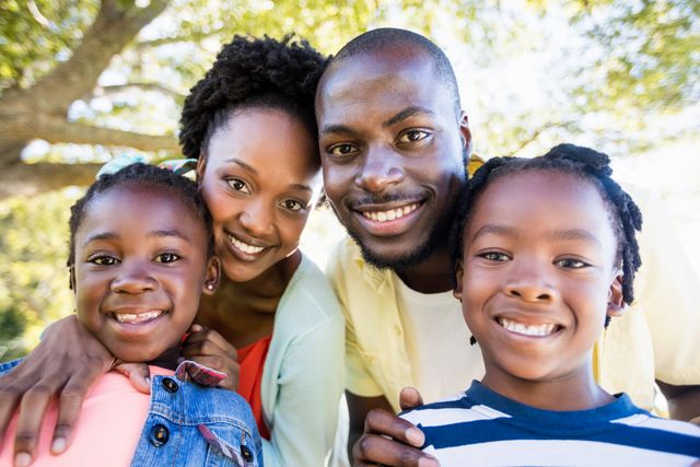 This image depicts a happy African American family enjoying time together in a park. The parents and two children are smiling warmly at the camera, radiating happiness and togetherness. Perfect for use in family-themed advertisements, brochures, magazines, or any content promoting family values, outdoor activities, and positive emotions.