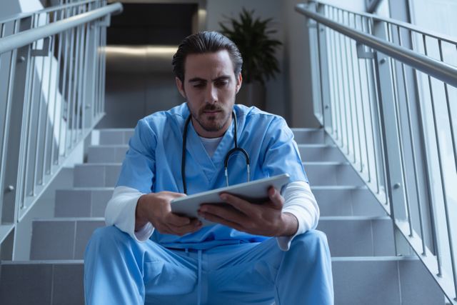 Male surgeon in scrubs sitting on stairs in hospital using digital tablet. Useful for healthcare technology, medical profession, hospital environment, and focused work themes.