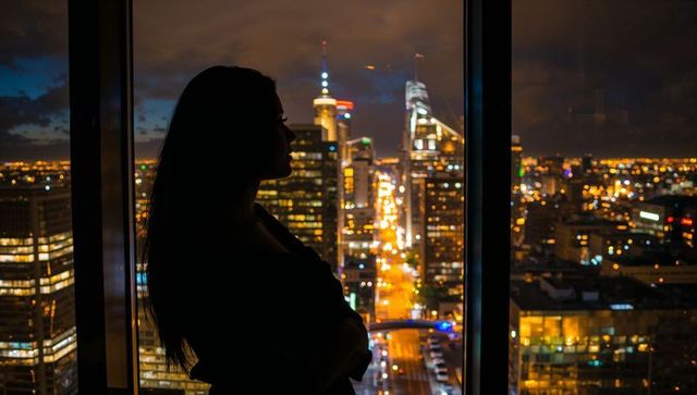 Silhouette woman gazing out high-rise window at night over vibrant urban skyline and city lights