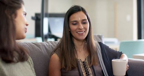 South asian woman wearing bindi chatting with friend on couch holding mug