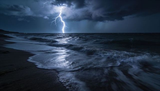 Branching lightning bolt illuminating night beach, stormy ocean waves and reflections