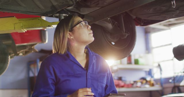 Female mechanic wearing blue overalls and safety goggles, inspecting car underside in auto repair workshop. Suitable for use in advertisements for automotive services, mechanic training programs, women's empowerment in technically-skilled jobs, and promotions for auto repair tools and equipment.