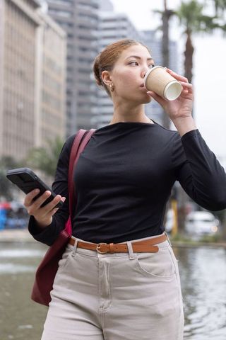 African american woman drinking coffee while checking smartphone in downtown urban plaza