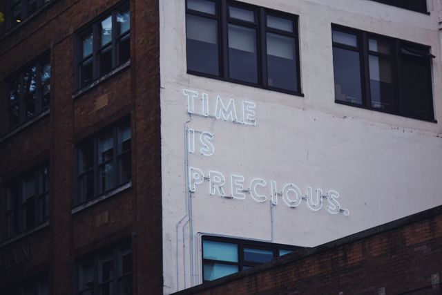 Neon sign on exterior of building displaying motivational message 'Time is Precious' with an urban background. Suitable for use in articles about city life, inspirational quotes, and urban art projects. Great for website banners, urban-themed designs, and social media posts focusing on the importance of time.