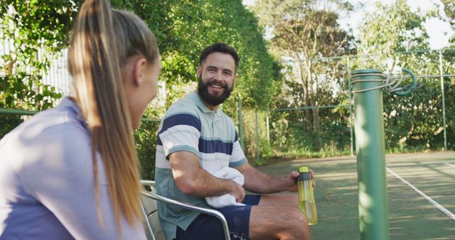 Two friends relaxing and chatting after a tennis match, sitting on a bench on a sunny day. Great for topics related to sportsmanship, friendship, outdoor activities, leisure, and hydration. Perfect for use in sports advertising, health and lifestyle blogs, or promotional materials for outdoor events.