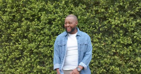 African american man standing smiling by green hedge wearing denim shirt and wristwatch