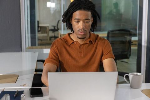 Focused african american man working on laptop in modern coworking space with coffee cup