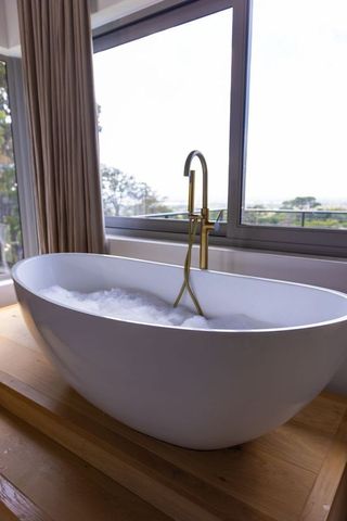 Minimalist freestanding bathtub with brass floor-mounted faucet and timber deck by window